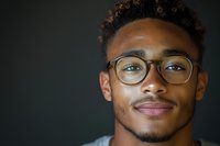 Young African American man wearing glasses smiles confidently at camera against dark background, showcasing natural curly hair and modern eyewear in professional headshot style.
