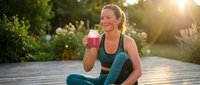 Young woman with blonde hair, Caucasian ethnicity, sitting outdoors on wooden deck holding pink smoothie drink after workout, surrounded by blooming flowers and green plants during golden hour sunset.