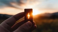 Hand holding battery icon with lightning bolt symbol against dramatic sunset sky, silhouetted fingers gripping smartphone screen displaying power indicator.