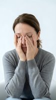 Middle-aged Caucasian woman in gray sweater covering face with hands, expressing stress, exhaustion, or emotional pain on light background.