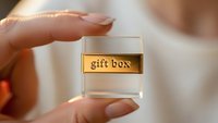 Hand holding small transparent gift box with golden label, luxury packaging concept for special occasions and premium presents.