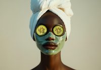 Young African woman wearing green facial mask and cucumber slices on eyes during spa skincare routine, wrapped in white towel against neutral background.