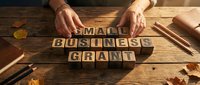 Entrepreneur hands arranging wooden letter blocks spelling small business grant on rustic desk with office supplies and autumn leaves for funding concept.