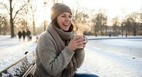Happy young Caucasian woman in knit sweater and beanie holding warm drink outdoors in snowy winter park during golden hour sunset with people in background.