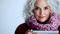 Mature Caucasian woman with silver curly hair wearing pink knitted scarf and burgundy sweater smiling warmly at camera against light background.