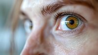 Close up macro shot of bright hazel eye with detailed iris pattern and long eyelashes against soft blurred skin background, showing natural eye structure.