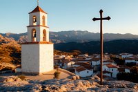 White church bell tower and wooden cross overlooking Mediterranean village with red tile roofs at sunset, mountains in background against blue sky.