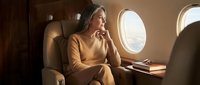 Young professional Caucasian businesswoman in beige suit working on laptop during luxury private jet flight, looking thoughtfully out airplane window.
