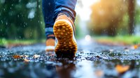 Closeup of yellow rubber boot sole stepping on wet asphalt during autumn rain, with fallen leaves and water droplets creating atmospheric outdoor scene.