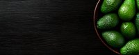 Fresh green avocados in dark wooden bowl on black wooden table background, top view with copy space for text. Food photography layout.