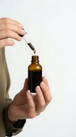 Woman hands using glass dropper to dispense essential oil or serum from amber bottle for skincare wellness routine on white background.