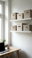 Minimalist home office with white floating shelves holding beige linen storage boxes, wooden desk, laptop, and mug near a bright window.