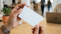 Close-up of hands holding blank white card in modern office workspace with laptop and potted plant visible in background.