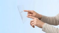 Hands holding transparent tablet computer with touchscreen interface against light blue background for modern technology concepts.