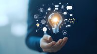 Glowing light bulb with business icons floating above open hand against blue background, representing innovation and digital transformation in business.