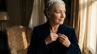 Elegant senior Caucasian woman adjusting black blazer in luxurious hotel room interior with warm lighting for business and lifestyle concepts.