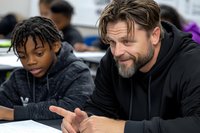 Caucasian male teacher with beard mentoring African American teenage student in classroom setting, showing supportive engagement during educational activity.