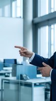 Male professional in dark business suit gesturing with both hands while presenting or explaining in modern office environment with glass partitions and natural light from windows.