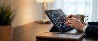 Professional businessman analyzing financial data on laptop computer in modern office workspace with warm lighting and plant decoration.