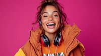 Young mixed race woman with bright smile wearing orange winter jacket and headphones on vibrant pink background, expressing joy and positive emotions.