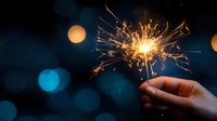 Glowing sparkler held in hand against dark background with blue and orange bokeh lights creates magical festive atmosphere. Celebration moment captured.