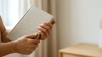 Person with medium skin tone holding closed beige linen book with both hands, wearing red nail polish, near window with natural light and neutral interior background.