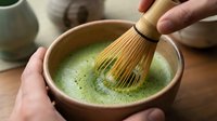 Hands whisking matcha powder in ceramic bowl with bamboo whisk, creating frothy green tea for traditional Japanese tea ceremony preparation.