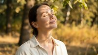 Young Asian woman with closed eyes enjoying warm sunlight in autumn park, peaceful moment of mindfulness and relaxation outdoors.