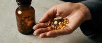 Male hand holding golden supplement capsules with amber bottle on neutral surface, representing wellness and nutritional health products.