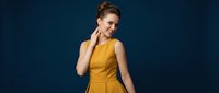 Young professional woman in yellow sleeveless dress touching neck, smiling confidently against dark blue background for business and lifestyle content.