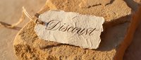 Vintage paper discount tag with elegant calligraphy script resting on rustic stone surface, warm sandy tones evoking savings and retail promotion concepts.