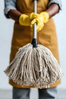 Close-up of person in yellow rubber gloves and brown apron holding cotton string mop against white background, representing cleaning service.
