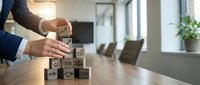 Professional businessman building wooden blocks with team icons on conference table in modern office boardroom representing teamwork and leadership development.