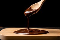 Melted dark chocolate dripping from wooden spoon onto bamboo surface against black background, creating glossy swirl pattern in dramatic lighting.