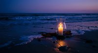 Glowing candle lantern on ocean beach at twilight with waves and purple sky creating romantic peaceful atmosphere for meditation and relaxation.