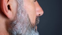Close up side view of mature Caucasian male with gray beard and ear against dark background, showing facial profile details and skin texture in soft lighting.