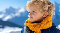 Young blonde boy in yellow knitted scarf enjoying winter mountain vacation with snowy peaks background for family travel and outdoor adventure content.