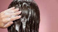 Woman applying shampoo to wet dark hair during washing routine against pink background for hair care and beauty concepts.