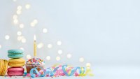 Colorful birthday celebration setup with macarons, cupcake with candle, party streamers and bokeh lights on white background for festive occasions.