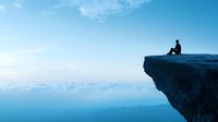 Silhouette of person sitting alone on cliff edge overlooking misty mountain landscape at dawn, contemplative moment in nature with blue morning sky.
