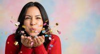 Young Hispanic woman in red sweater blowing colorful confetti from hands against pastel rainbow background for celebration and party concepts.