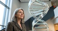 Mature Caucasian female scientist in professional attire standing beside large DNA double helix model in modern laboratory or research facility.