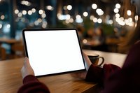 Person using tablet with blank white screen in cozy cafe setting with warm bokeh lights and coffee cup on wooden table for mockup presentations.