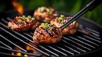 Juicy glazed chicken thighs with sesame seeds and green onions grilling on barbecue grate. Tongs turning meat against flames and smoke background.