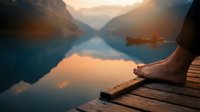 Person relaxing by serene mountain lake at sunset with perfect reflections on calm water surface creating peaceful meditation moment.
