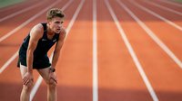 Caucasian male athlete in black sleeveless top catching breath on orange running track with white lane markings, displaying exhaustion after sprint training or competition.