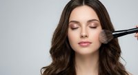 Professional makeup artist applying blush to young Caucasian woman with closed eyes and wavy brown hair using cosmetic brush on neutral background.