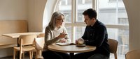 Young couple enjoying coffee date conversation at modern cafe table by large window with natural lighting and minimalist interior design.