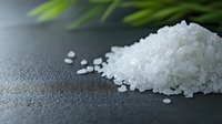 White sea salt crystals scattered on dark stone surface with soft green plant blur in background, macro food photography for culinary and wellness concepts.