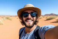 Happy bearded man taking selfie in desert landscape wearing straw hat and sunglasses during adventure travel vacation under clear blue sky.